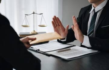 A man in a black suit and striped tie raises both hands in refusal as another person slides an envelope across a desk with legal documents and a gold balance scale in the background depicting an attorney from Rueb Stoller Daniel advising a client to reject a lowball first settlement offer from an insurance company in California.